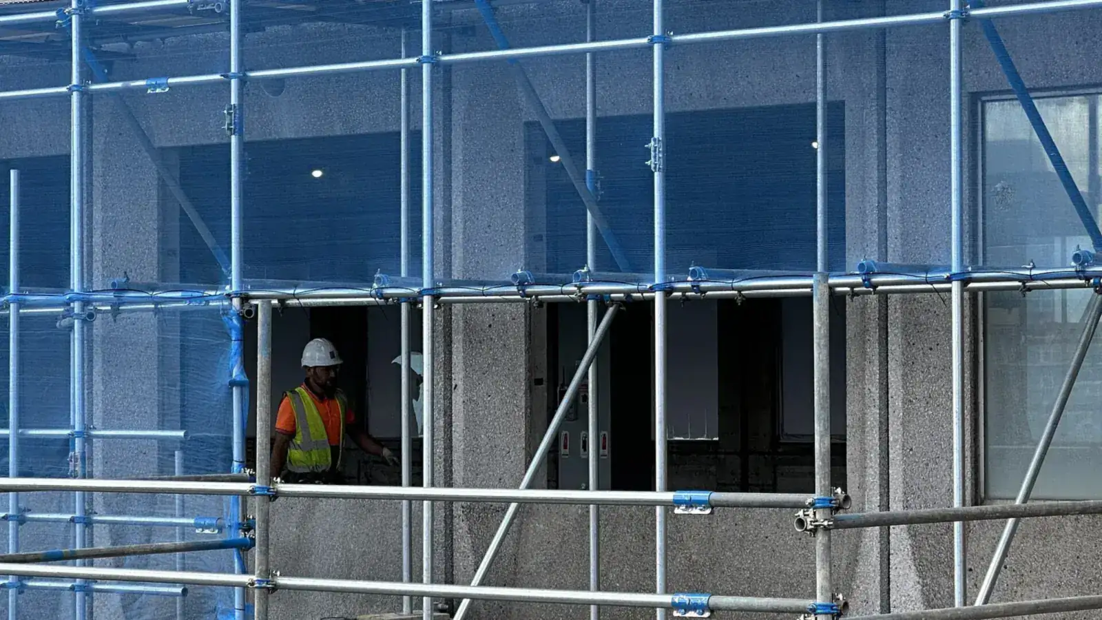 Construction worker in a hard hat and high-visibility vest on a tube-and-coupler scaffold beside a building
